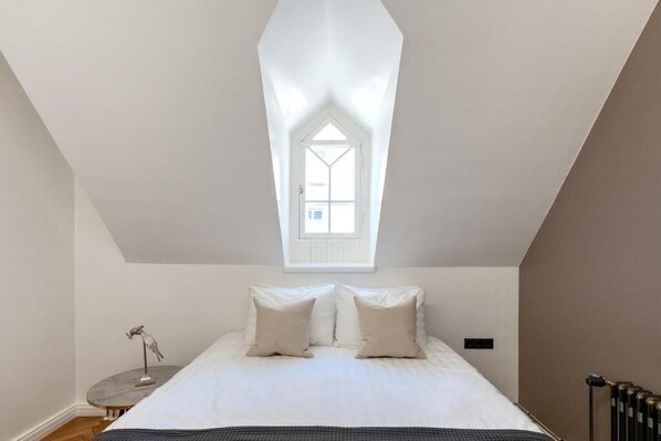 Un dormitorio con tragaluz, una cama con ropa blanca, una mesita de noche pequeña con una lámpara y un radiador.
