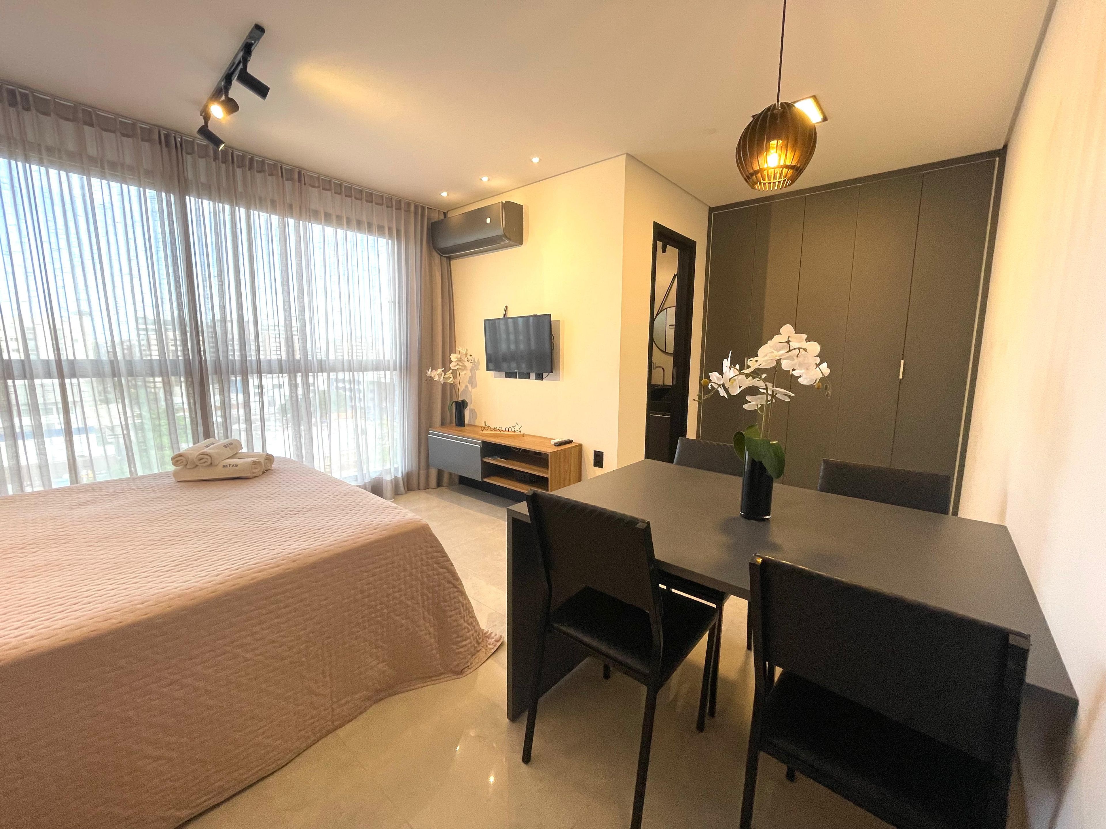 Habitación de hotel moderna con cama, una mesa de comedor con sillas, televisión y un ventanal con cortinas.