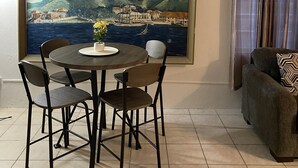 Un comedor con una mesa redonda y cuatro sillas, un cuadro de una escena costera en la pared, un aire acondicionado y un sofá con un cojín estampado.