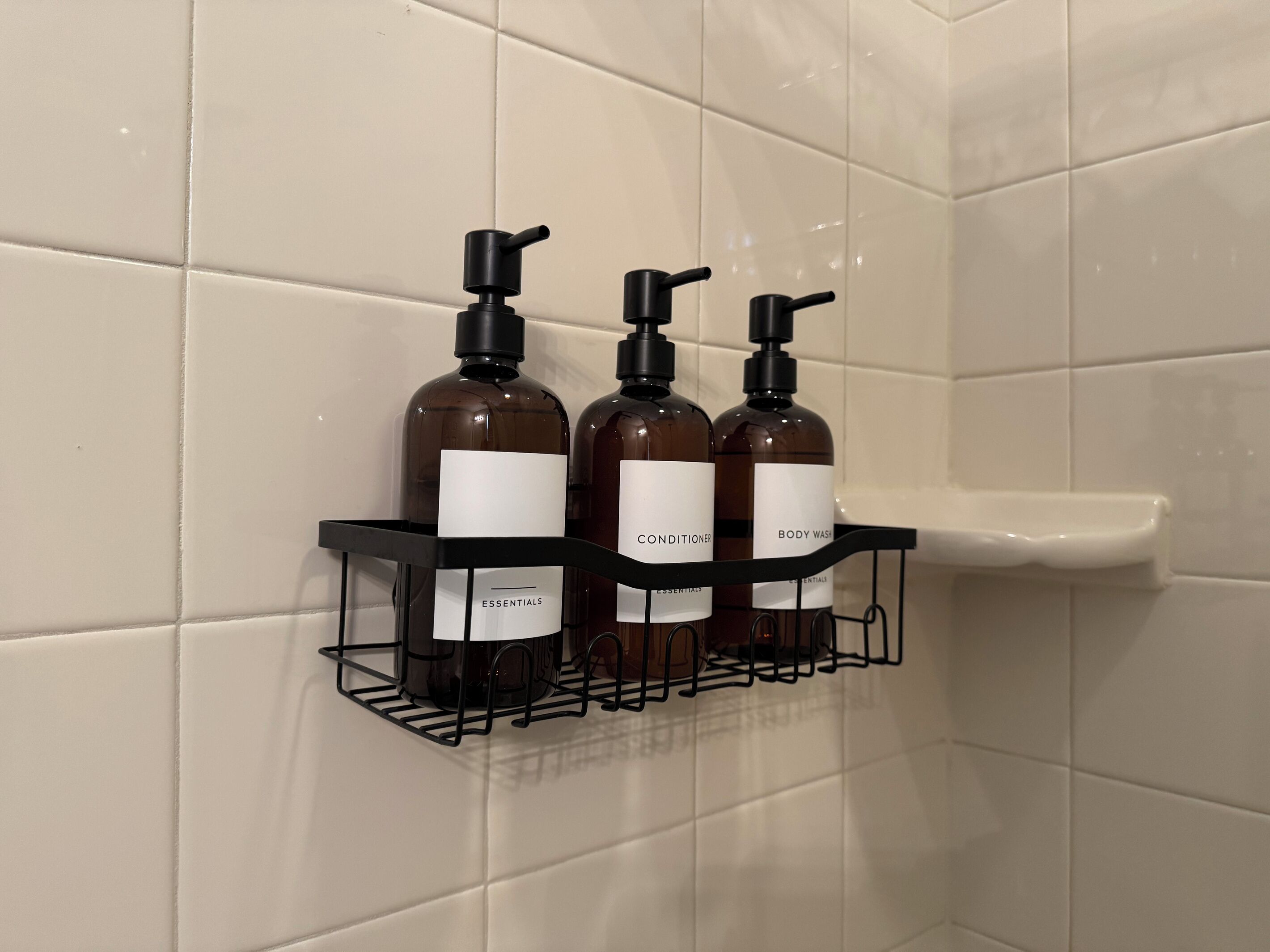 Een badkamer met een betegelde wand en een zwart schap met drie flessen toiletartikelen.