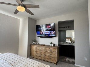 Une chambre à coucher comprenant un lit, une commode en bois, un téléviseur fixé au mur et un ventilateur de plafond.