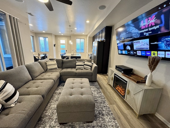 A modern living room with a sectional sofa, ottoman, fireplace, flat-screen TV, and a dining area in the background.