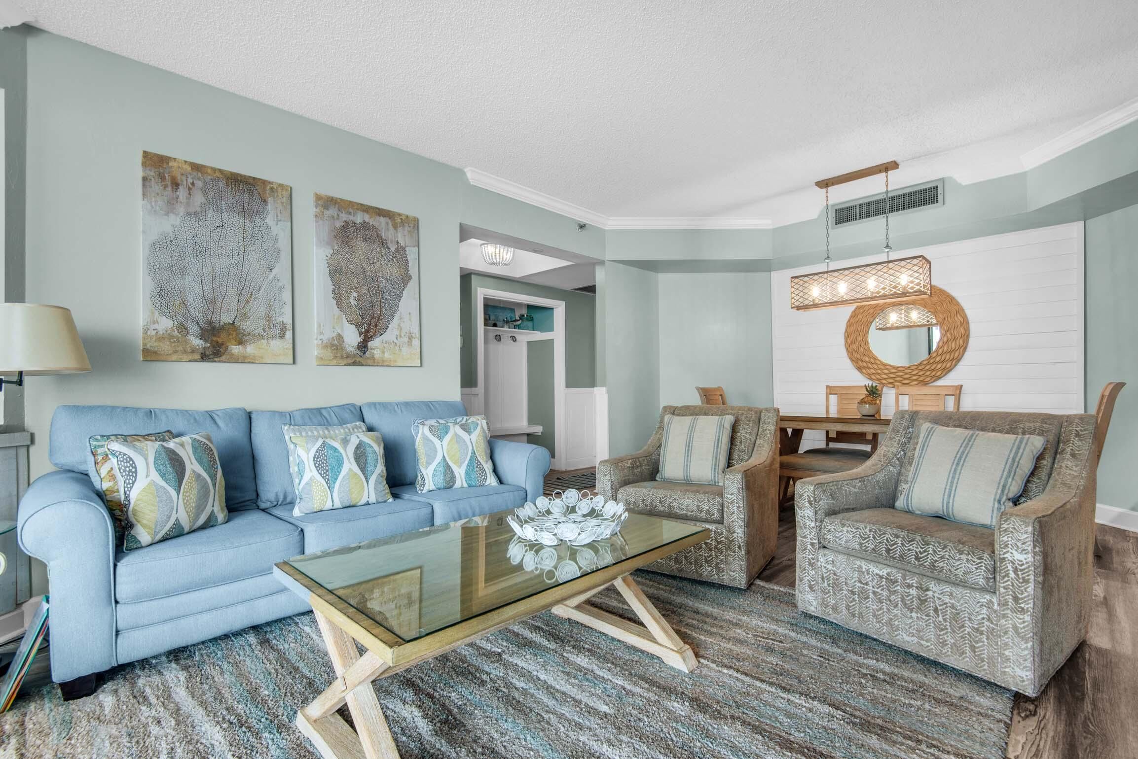 A living room with a blue sofa, patterned armchairs, a glass coffee table, and abstract wall art.