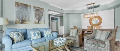 A living room with a blue sofa, patterned armchairs, a glass coffee table, and abstract wall art.