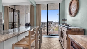 A modern kitchen with a marble countertop, wooden chairs, and a view of the ocean through large sliding doors. - Surfside Resort #404 (Miramar Beach)