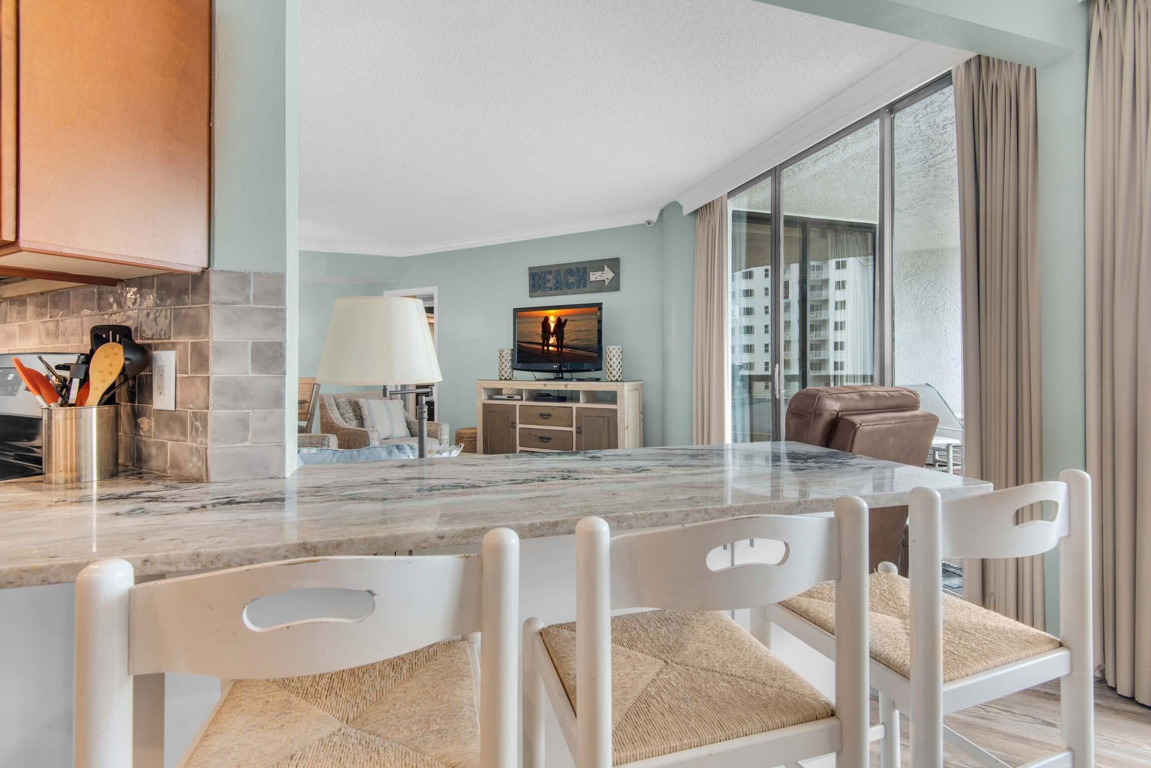 A modern kitchen with a marble countertop, white chairs, and a television displaying a beach scene.