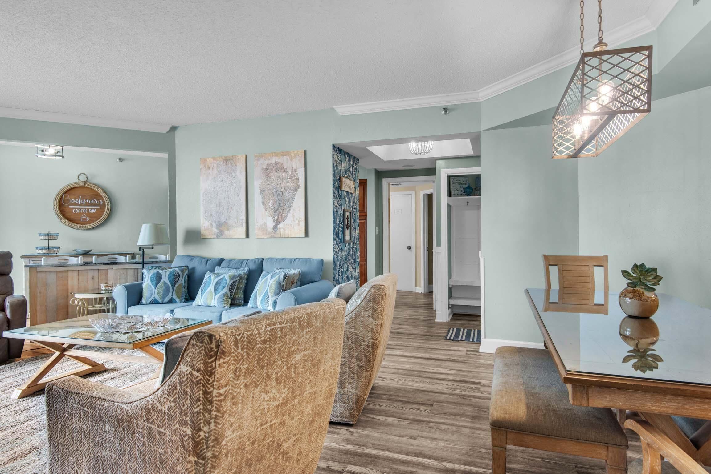 Ein modernes Wohnzimmer mit einem blauen Sofa, gemusterten Sesseln, einem Glastisch und einem Essbereich mit Holztisch und Stühlen.