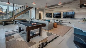 A spacious living area with a pool table, comfortable seating, a staircase, and a large TV wall.