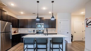 A modern kitchen with dark cabinets, stainless steel appliances, and a central island with bar stools. - Landing | Modern 2BD, Clubhouse, Yoga Studio (Buford)