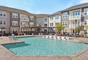 A pool area with lounge chairs, a pergola, and a multi-story building with balconies. - Landing | Beautiful 2BD, Pool, Game Room (Matthews)
