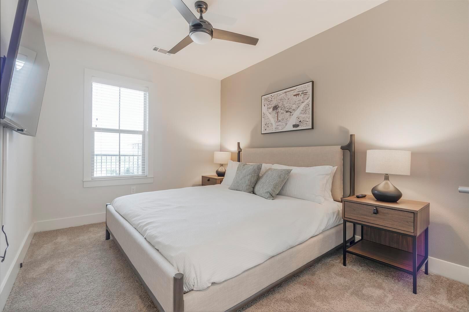 A bedroom with a bed, two bedside tables, a ceiling fan, a window, and a wall-mounted artwork.