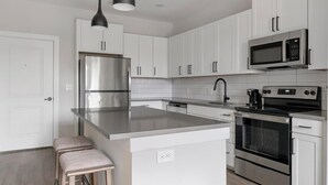 Una cocina moderna con armarios blancos, electrodomésticos de acero inoxidable, una isla grande con taburetes y una lámpara colgante negra.