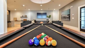 Una mesa de billar con bolas y tacos, un salón moderno con sofás, un televisor y un cuadro en la pared.