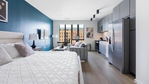 A modern hotel room with a large bed, a kitchenette, and a view of buildings through the windows. - Landing | Stylish Studio, Rooftop Lounge, Pool (Durham)