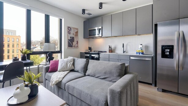 A modern kitchen with grey cabinets, a stainless steel refrigerator, and a microwave. There is a dining area with a grey sofa and a table with a plant. - Landing | Stylish Studio, Rooftop Lounge, Pool (Durham)