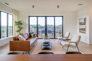 A modern living room with a leather sofa, a coffee table with books, and a view of a balcony with outdoor furniture. - Landing | Spectacular 2BD, Gym, Rooftop Lounge (Durham)