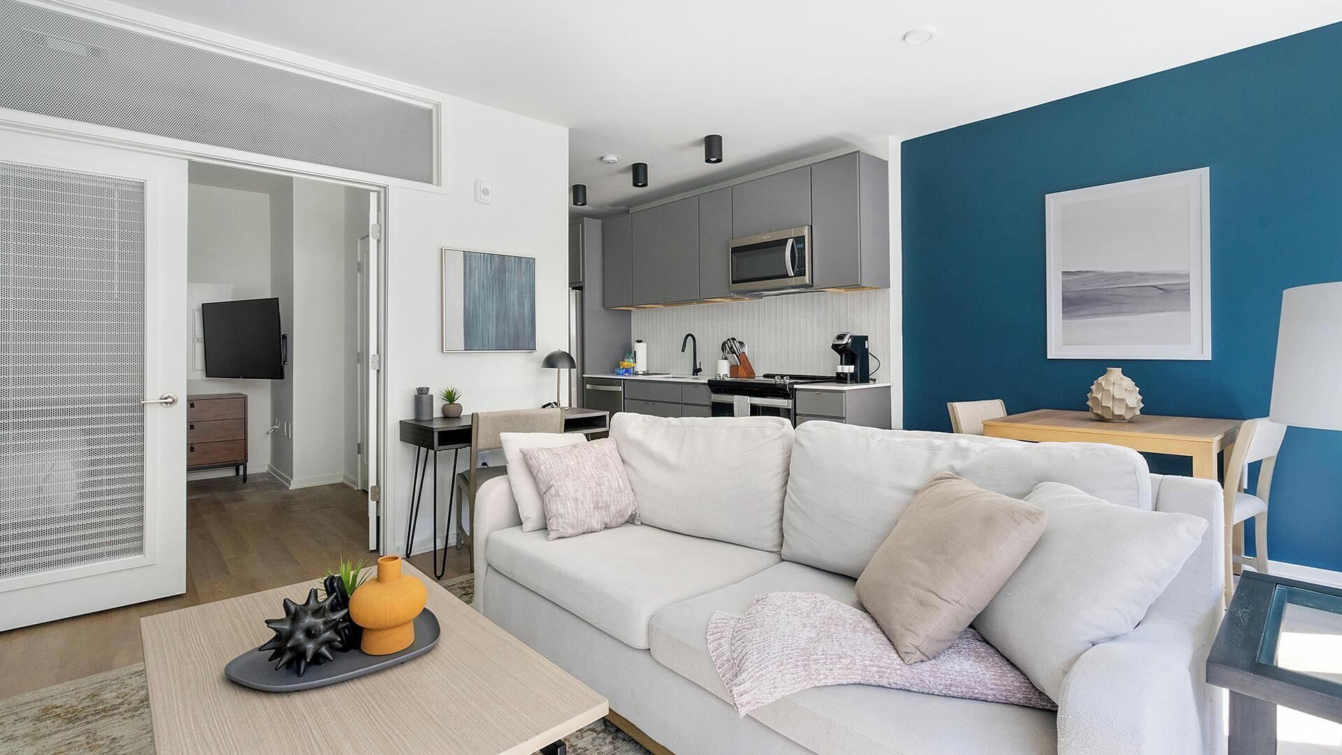 Una sala moderna con un sofá blanco, una mesa de centro con objetos decorativos, una cocina al fondo con armarios grises y un microondas, y una pared azul de acento.