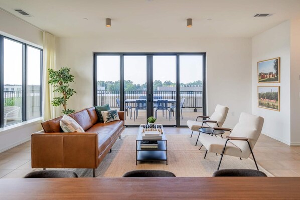 A modern living room with a leather sofa, a coffee table with books, and a view of a balcony with outdoor furniture.