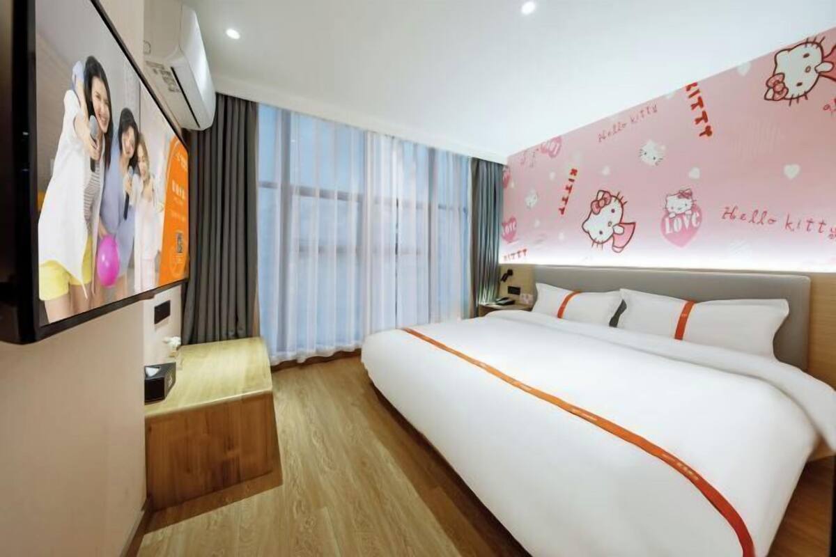 A hotel room with a large bed, a desk, a TV, and a wall with Hello Kitty decorations.