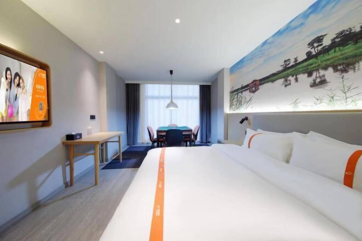 A hotel room with a large bed, a desk, a chair, a dining table, and a painting on the wall.