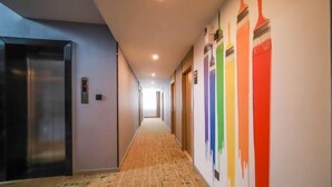 Un couloir d’hôtel avec un plancher en bois, un ascenseur métallique et un mur orné d’une fresque abstraite aux couleurs vives.