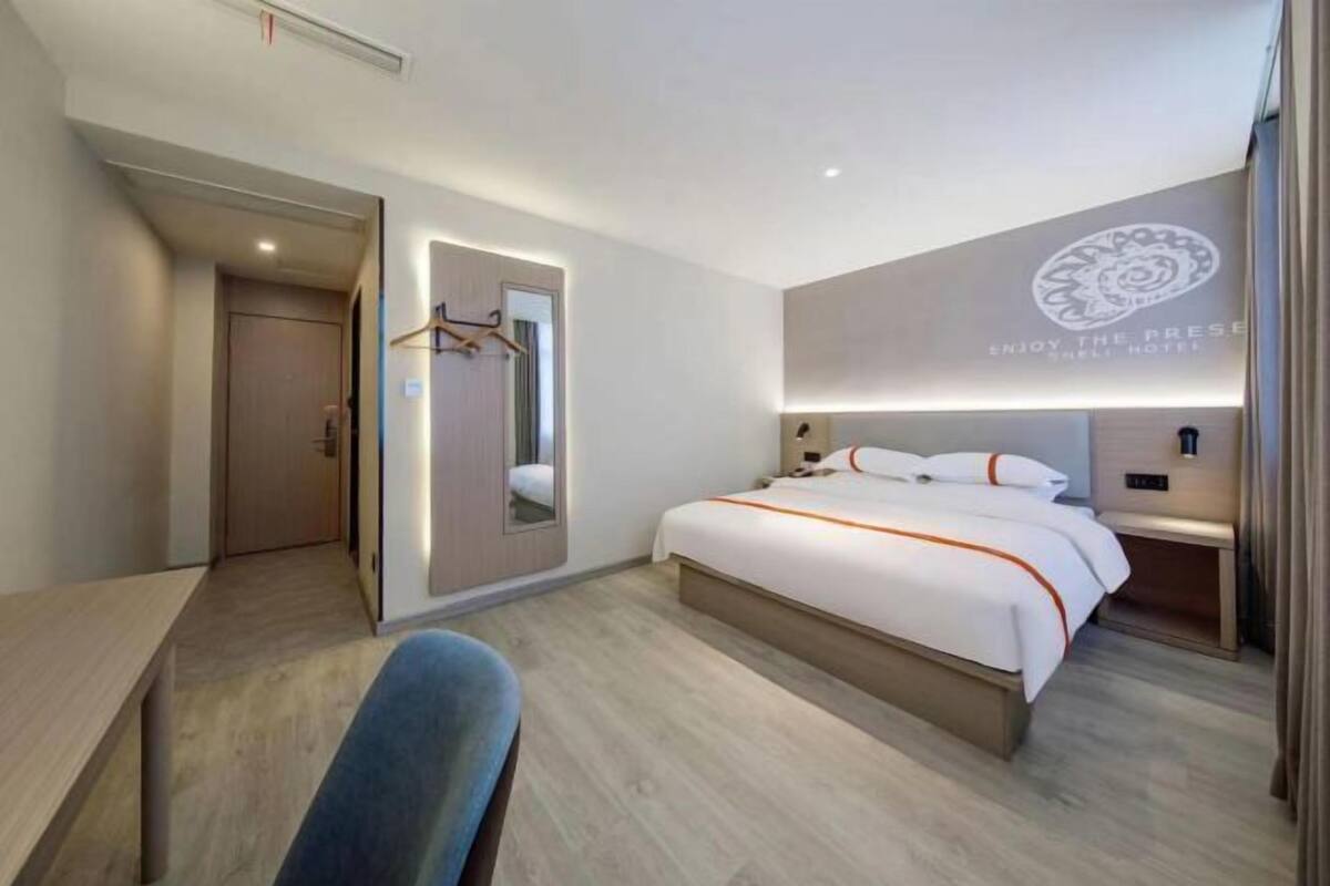A modern hotel room with a large bed, a desk, a chair, a wardrobe, and a mirror.