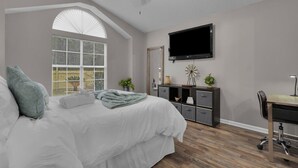 Ein Schlafzimmer mit Bett, Schreibtisch, Stuhl, Fernseher, Deckenventilator und einem Fenster mit Jalousien.
