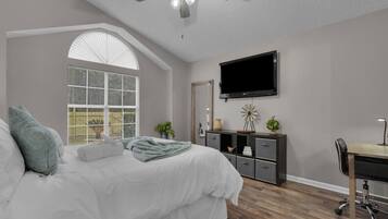 A bedroom with a bed, a desk, a chair, a TV, a ceiling fan, and a window with blinds.