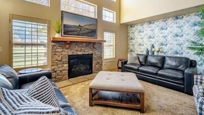 A living room with a stone fireplace, a black leather sofa, a tufted ottoman, and a large window with blinds. - Elegant Retreat & Small Gathering Home-Waffles too (Orem)