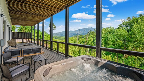 A wooden deck with outdoor seating and a hot tub, overlooking a forested landscape with mountains.