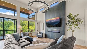 A modern living room with a large leather sofa, a flat-screen TV mounted on a wall, and a view of the outdoors through large windows.