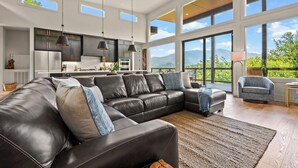 A spacious living room with a large leather sectional sofa, a kitchen with dark cabinets and stainless steel appliances, and large windows offering a mountain view.