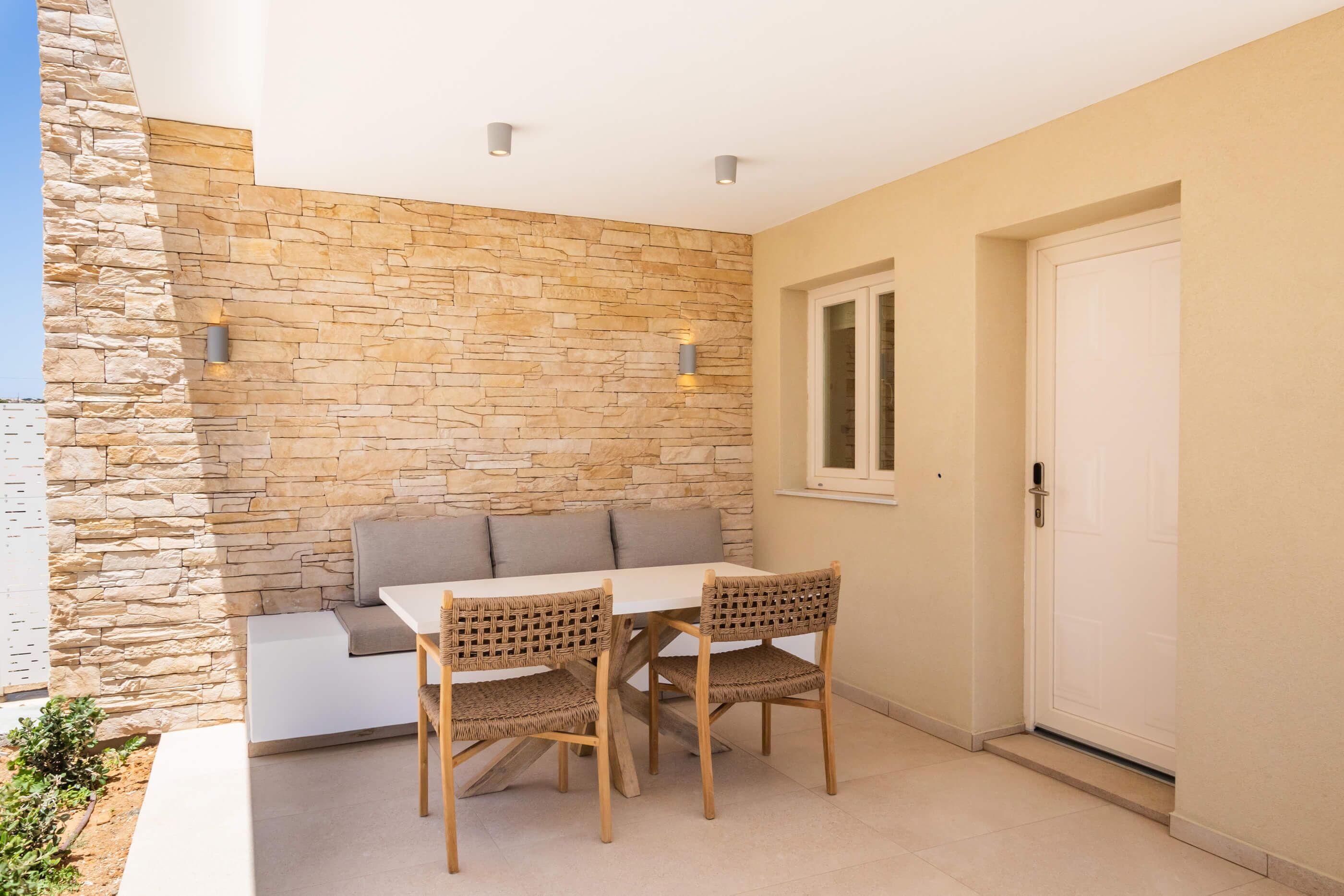 A patio area with a stone wall, a white table, two wicker chairs, and a grey sofa.