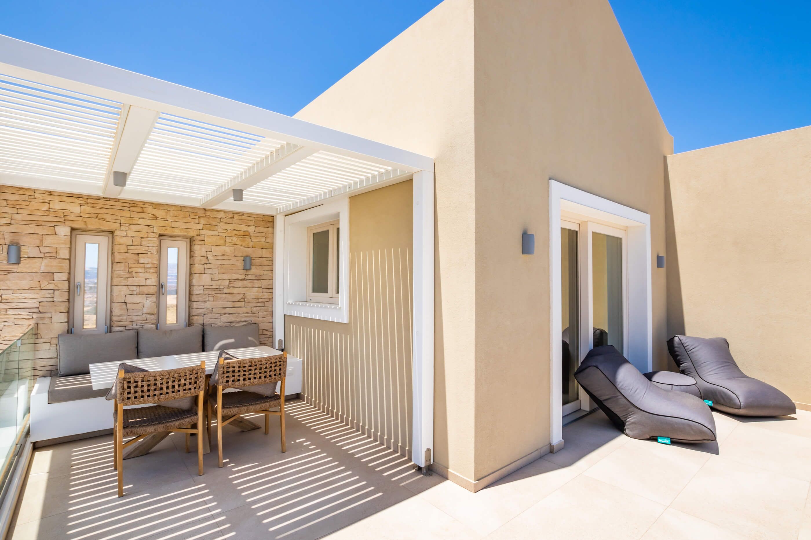 A modern outdoor patio with a stone wall, a seating area with cushions, a table, and two reclining chairs.