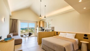 A modern hotel room with a large bed, a sofa, a small table, and a view of the outdoors. - Thalassa Suites (Karpathos)