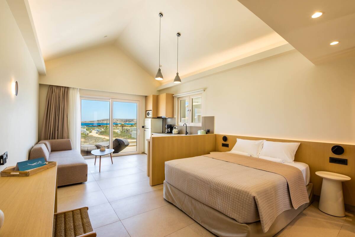 A modern hotel room with a large bed, a sofa, a small table, and a view of the outdoors.