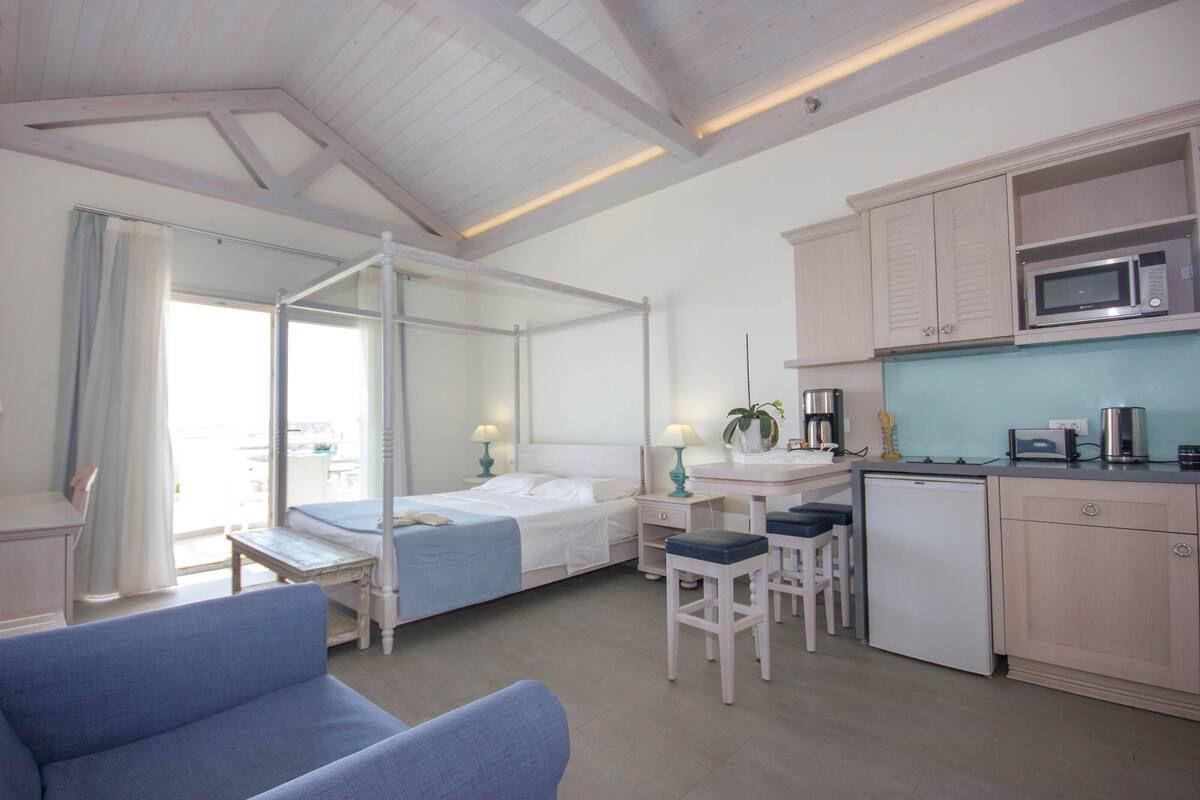 A hotel room with a bed, a desk, a kitchenette, and a balcony.