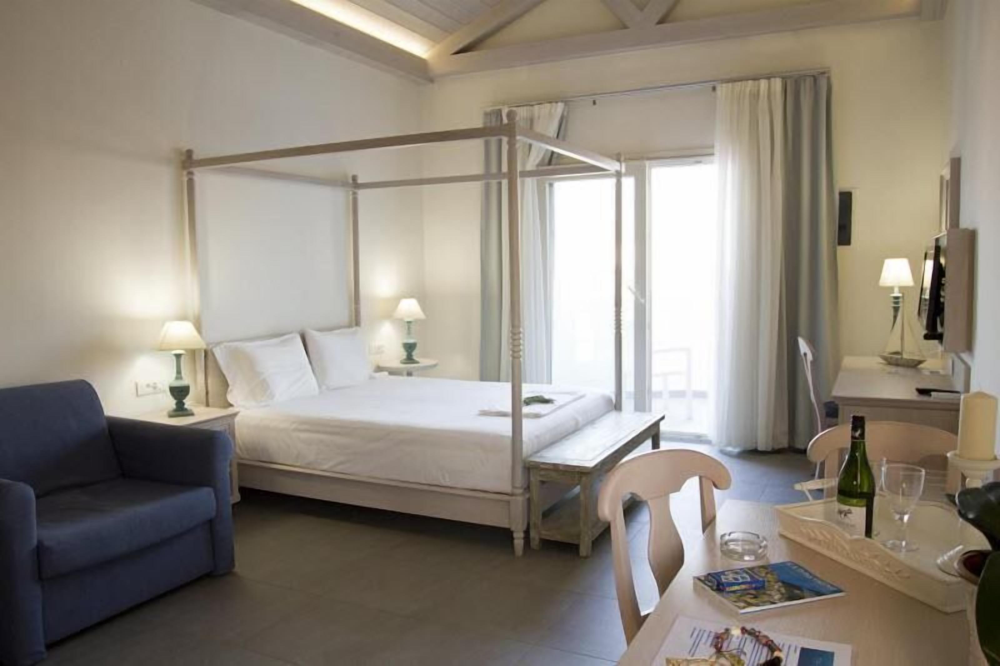 Una cama con dosel, ropa de cama blanca, una mesita de noche con lámpara, una silla y una mesa de comedor con una botella de vino y copas.