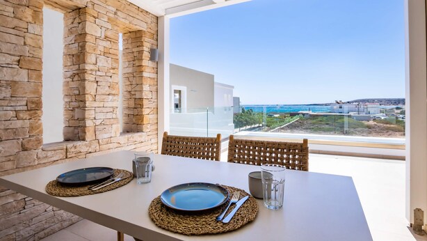 A dining table set with plates, cutlery, and glasses, overlooking a scenic view of the sea and buildings. - Thalassa Suites (Karpathos)