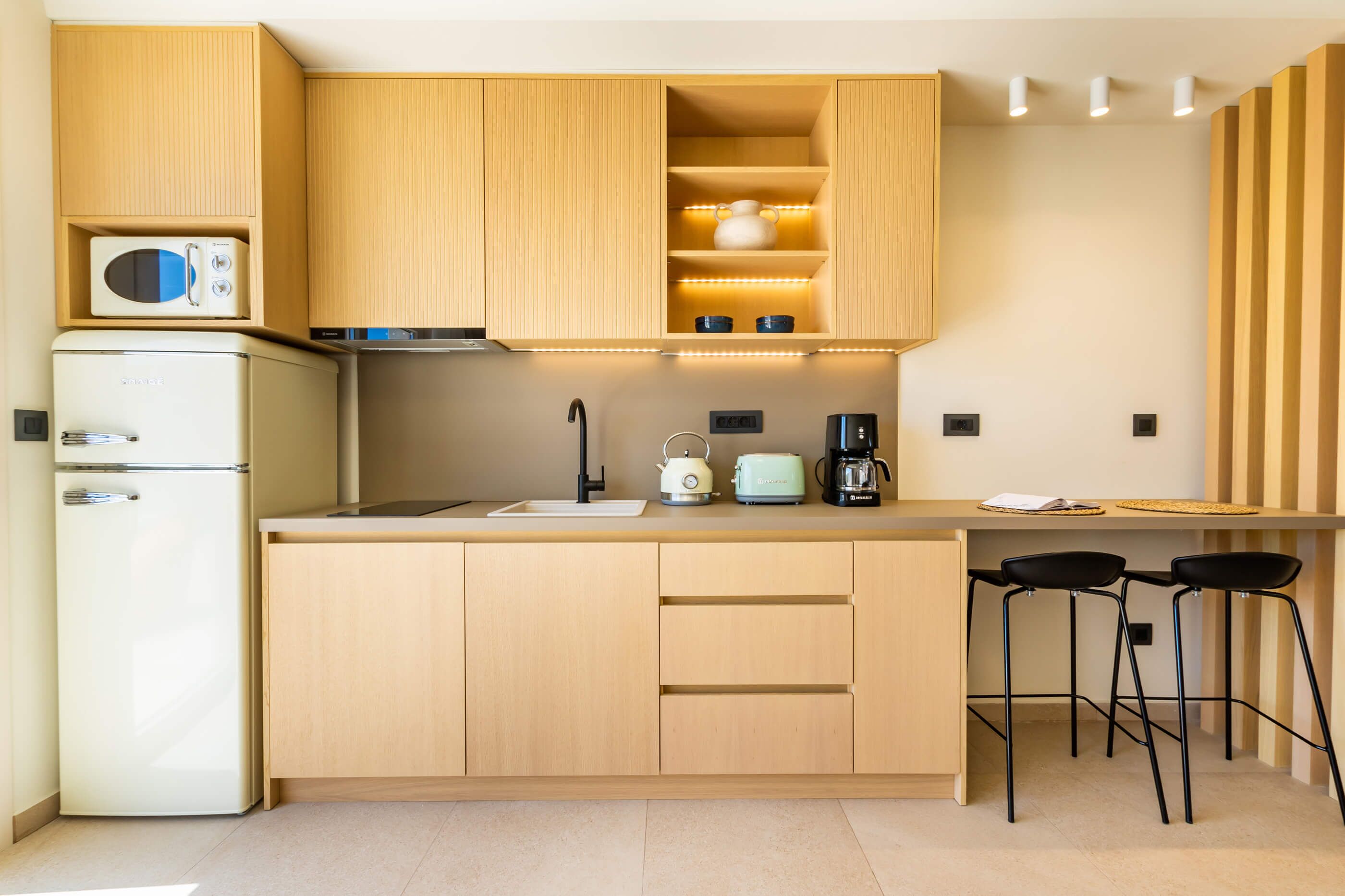 A modern kitchen with wooden cabinets, a built-in microwave, a coffee maker, and two bar stools.