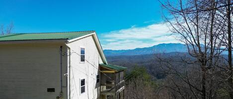 A white building with a green roof, a porch, and a view of mountains in the background.