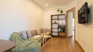 Un salon moderne avec un plancher en bois, un canapé vert, une petite table sur laquelle se trouve un verre de jus d’orange et une bibliothèque intégrée agrémentée de plantes.