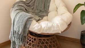 Un fauteuil rond et moelleux, doté d’une assise tissée, avec un plaid gris et une plante en pot à côté.