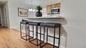 Une cuisine avec un plancher en bois, un mur blanc, un bar avec des tabourets en métal noir, une plante en pot et un four à micro-ondes.
