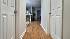 Un couloir au plancher en bois et aux portes blanches, qui mène à une pièce dot ée d’une cheminée et d’une plante.