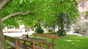 Une table de pique-nique en bois avec des bancs, une bouteille de vin et un verre de vin rouge, le tout installé dans un espace extérieur verdoyant, agrémenté d’arbres et avec un bâtiment en arrière-plan.