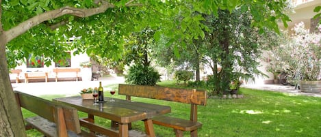 Un tavolo da picnic in legno con panche, una bottiglia di vino e un bicchiere di vino rosso, immerso in una zona erbosa con alberi e un edificio sullo sfondo.