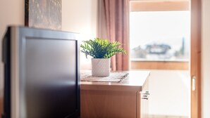 Um quarto com televisão sobre um suporte de madeira, uma pequena planta em vaso sobre uma mesa e vista para a cidade através de uma porta aberta.