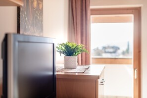 Ein Zimmer mit Fernseher auf einem Holzständer, einer kleinen Topfpflanze auf einem Tisch und Blick auf eine Stadt durch eine geöffnete Tür.