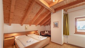Quarto com teto de madeira, cama com roupa de cama branca, mesa de cabeceira, janela com cortinas e radiador.